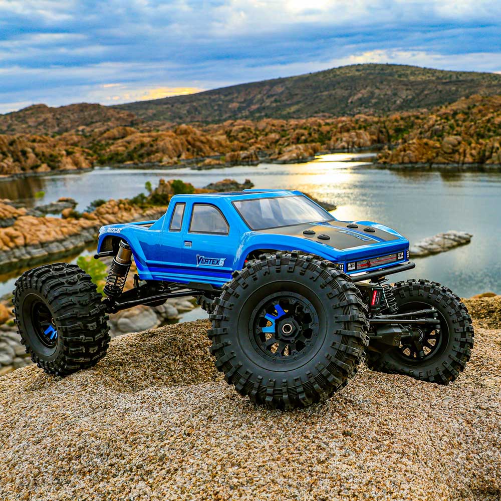 Blue off-road model truck on a rocky surface with a scenic background of water and mountains.