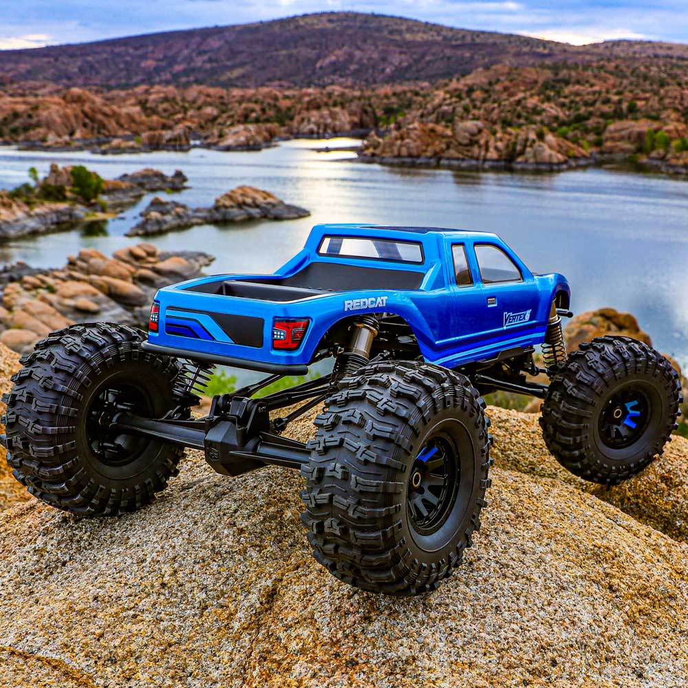 Blue remote-controlled monster truck on rocky terrain with a lake and mountains in the background