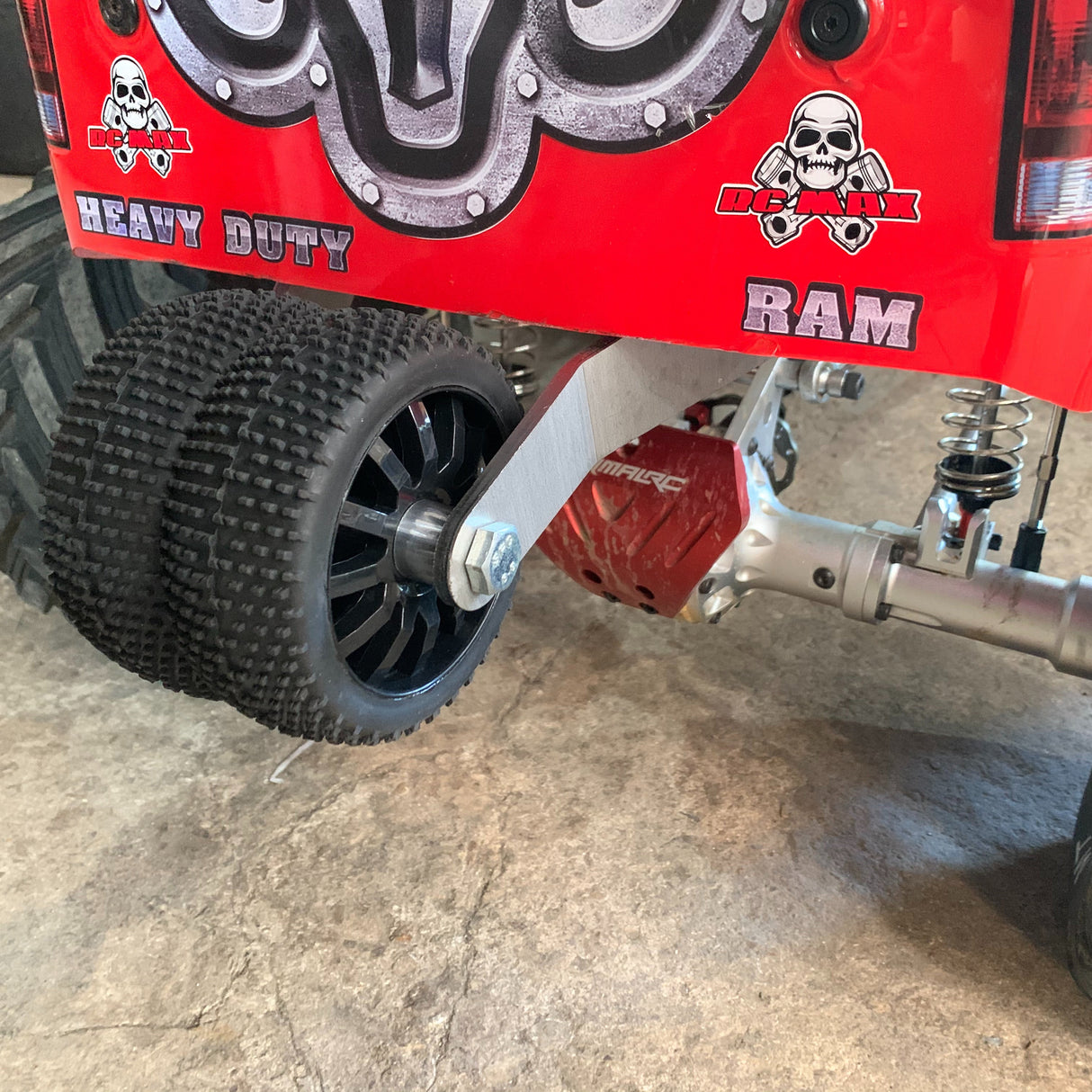 Close-up of a red RC crawler truck's rear wheel and suspension, featuring a black knobby tire, silver shock absorber, and a red brake caliper. The red body has 'HEAVY DUTY' and 'RAM' graphics with skull logos.